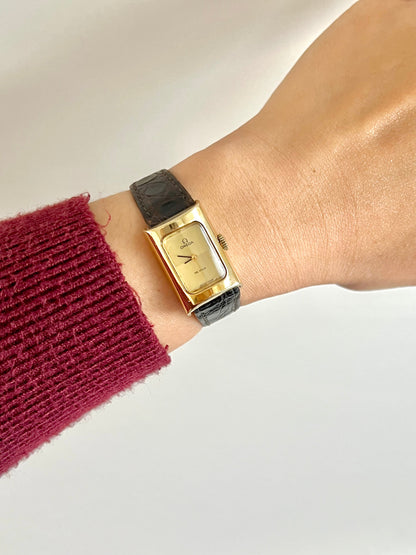 Omega De Ville gold rectangular watch with black strap shown on a wrist.