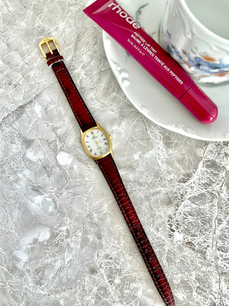 Omega De Ville women’s gold-tone oval watch with red strap laid flat in a styled photograph