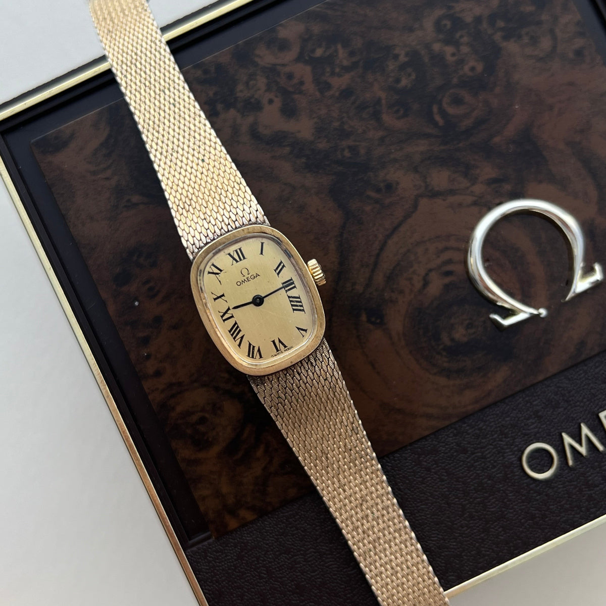 Vintage Omega De Ville women's watch featuring a champagne Roman numeral dial, rounded rectangular gold tone case and woven mesh bracelet displayed on an Omega presentation box