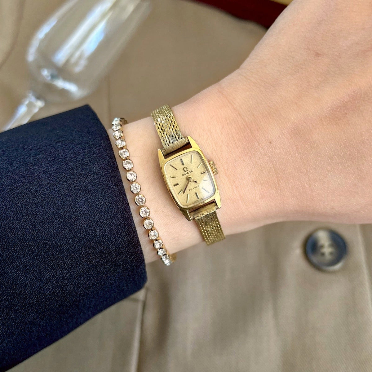 Omega De Ville women’s cocktail watch with a gold snowflake dial and rolled gold bracelet, worn beside a crystal bracelet