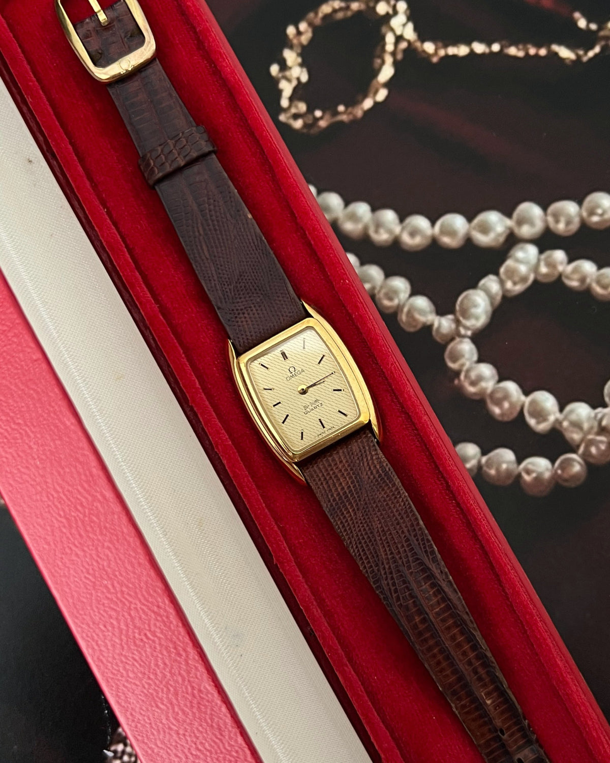 Omega De Ville watch with a wavy champagne dial, gold tone tonneau case and brown lizard strap in its red Omega box.