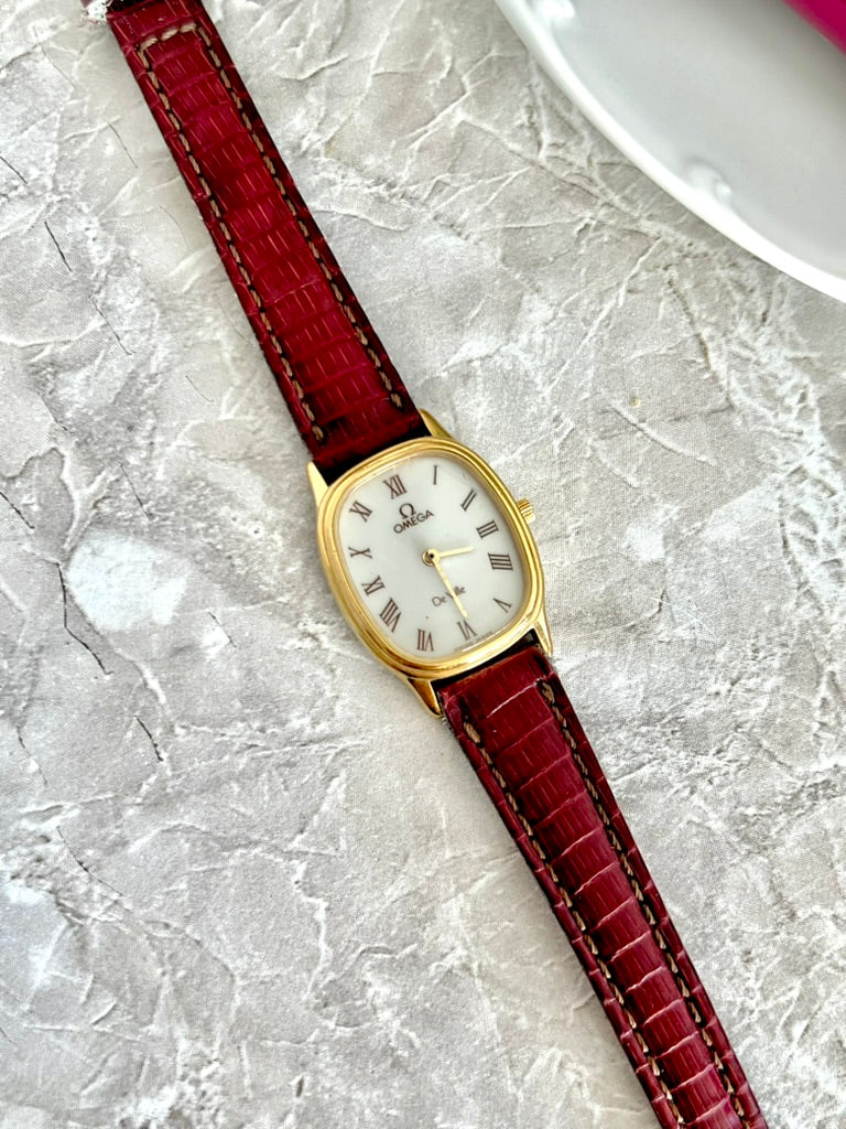 Close-up of Omega De Ville white dial, black Roman numerals, gold-tone oval case and red textured strap