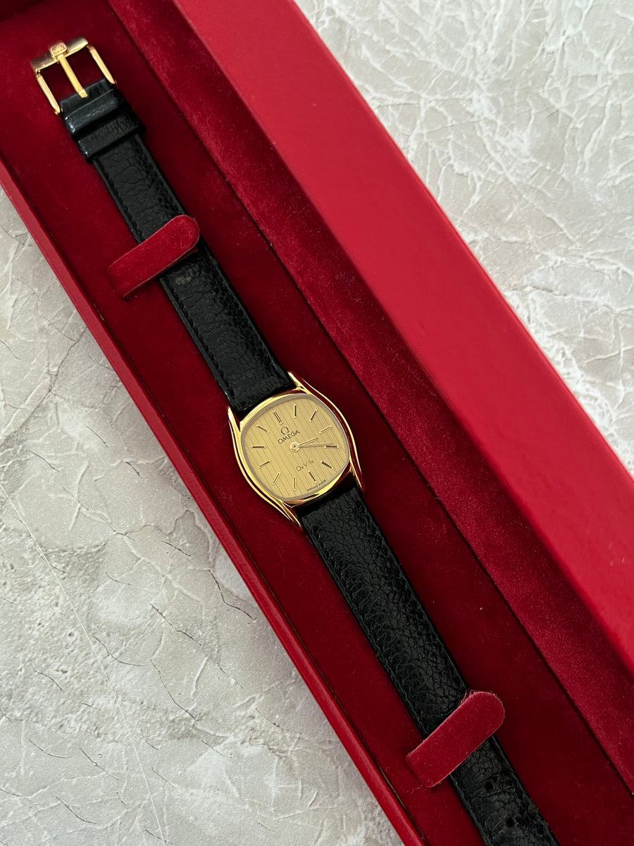 Omega De Ville women's gold-tone cushion watch with black leather strap and gold-tone buckle, photographed in a red box