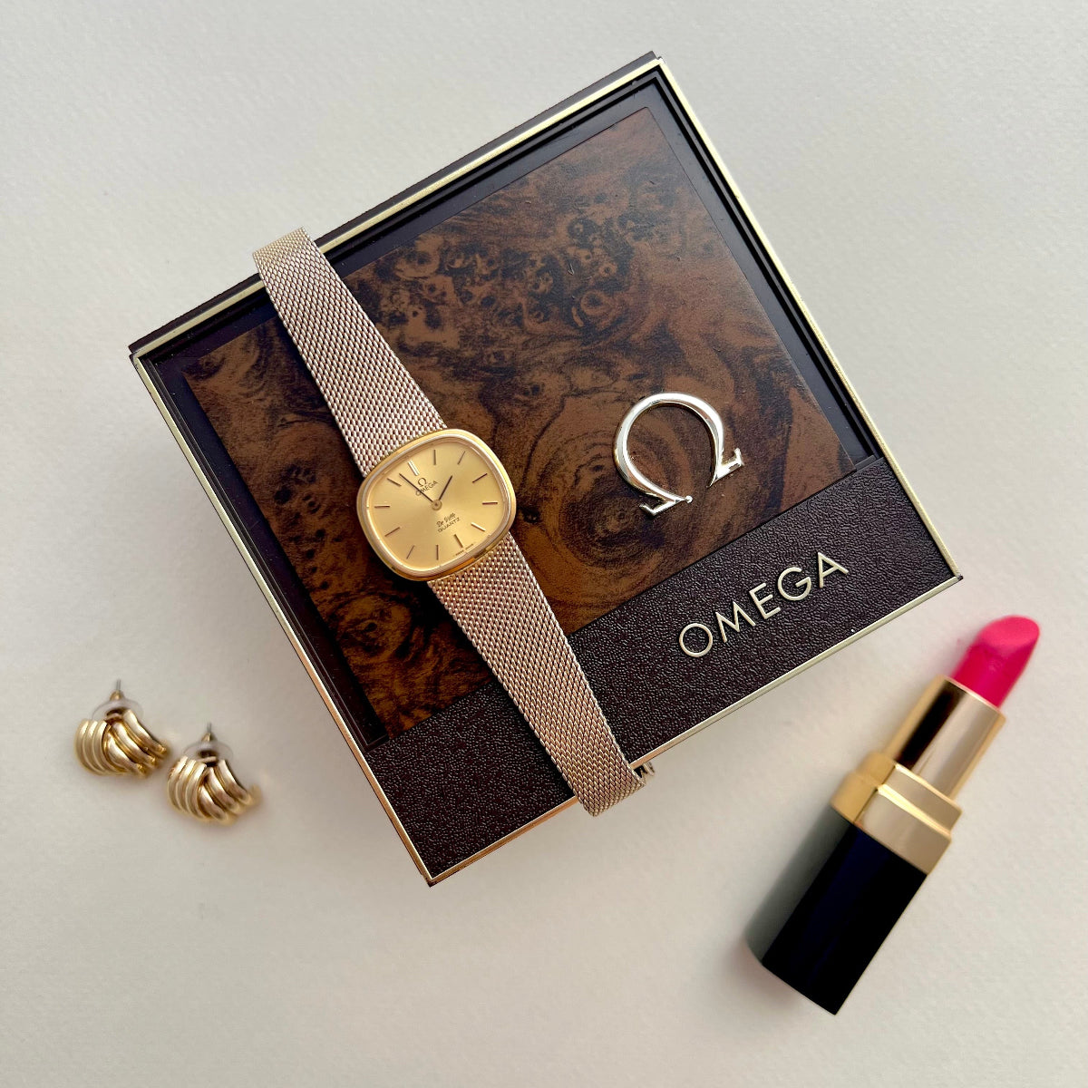Omega De Ville women's watch with a gold dial and Milanese mesh bracelet styled with Omega logo, gold earrings, silver bangle and pink lipstick in an elegant flat lay.