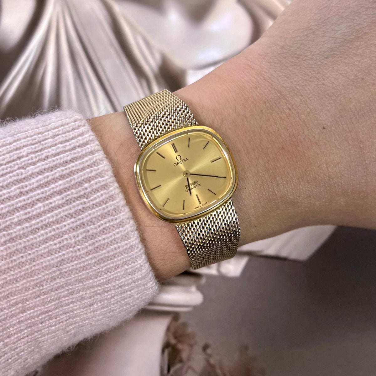 Omega De Ville women’s watch with gold tone mesh bracelet, champagne dial and softly rounded square case worn on wrist.