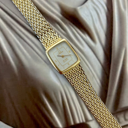 Omega De Ville gold women’s bracelet watch with square cream dial, Roman numeral accents and wide woven gold tone bracelet.