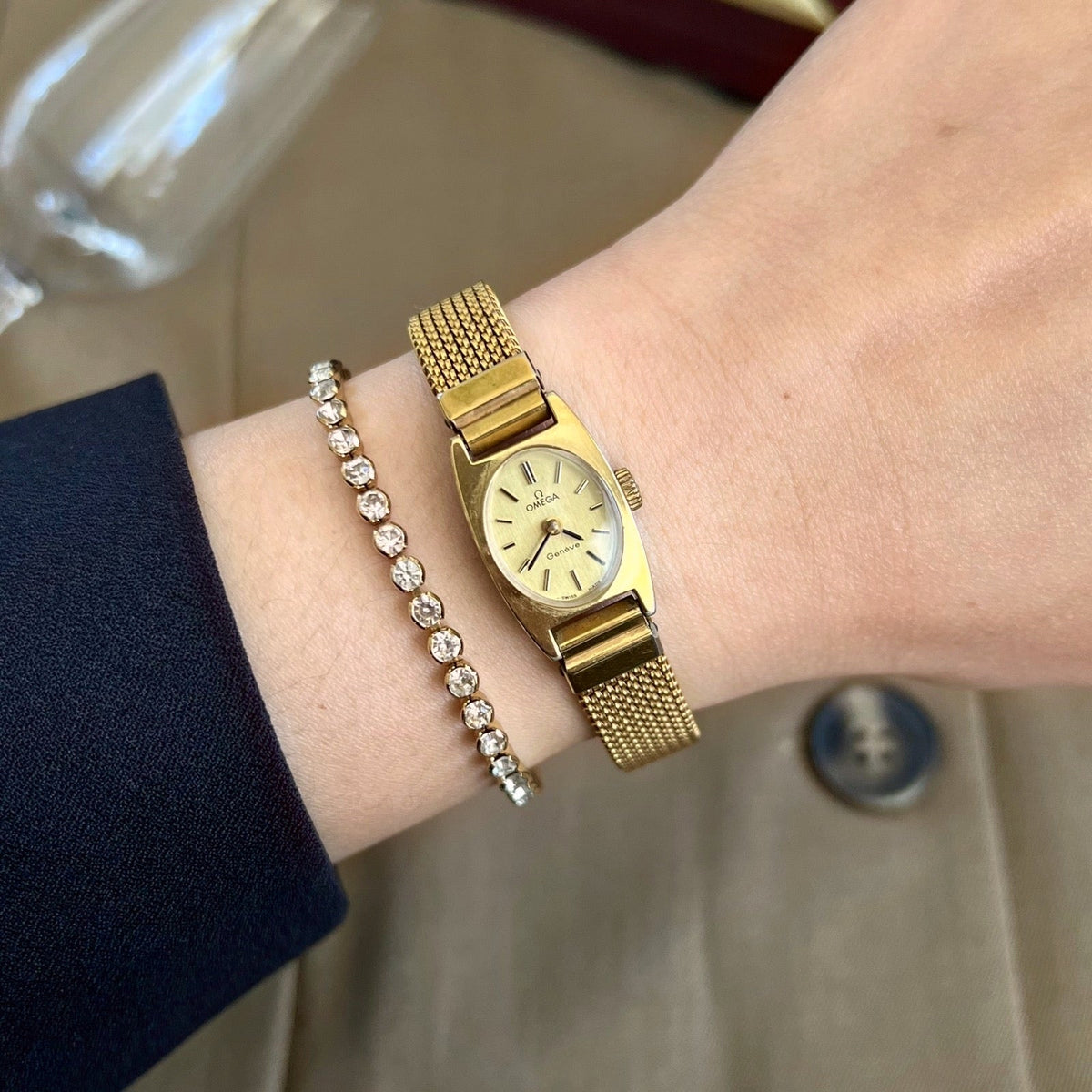Omega Geneve women’s watch with an oval champagne dial and gold mesh bracelet, worn beside a crystal bracelet