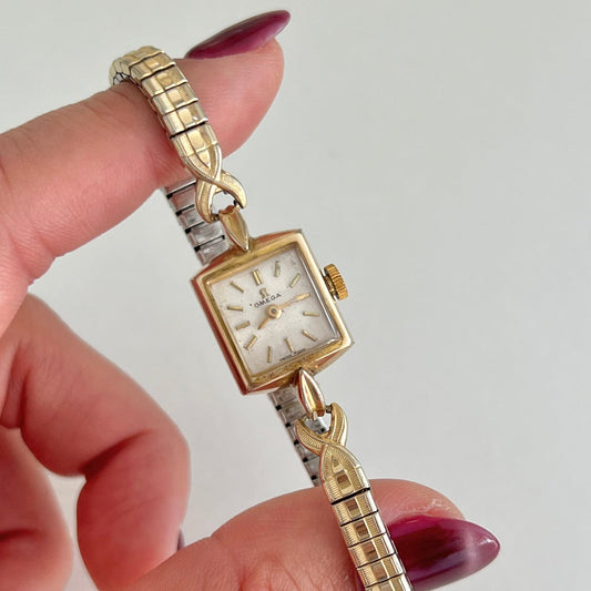 Omega Gold and silver watch held by a hand with purple nail polish on a light gray background