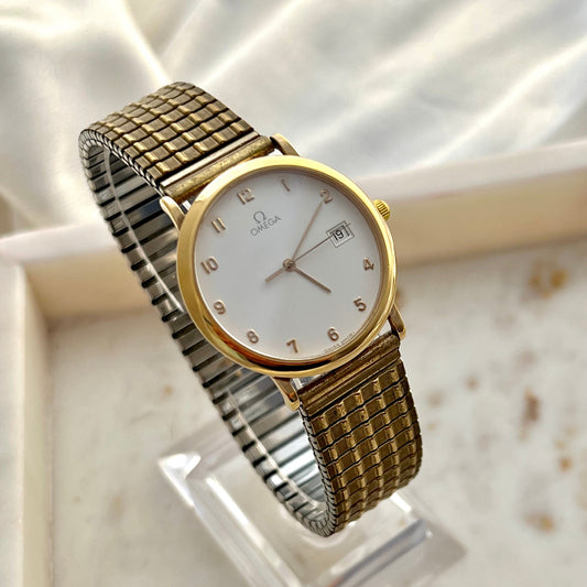 Omega women’s dress watch with white dial, gold tone round case, applied Arabic numerals, date window and woven jewellery style bracelet, elegant minimalist Swiss watch photographed by The Gilded Hour