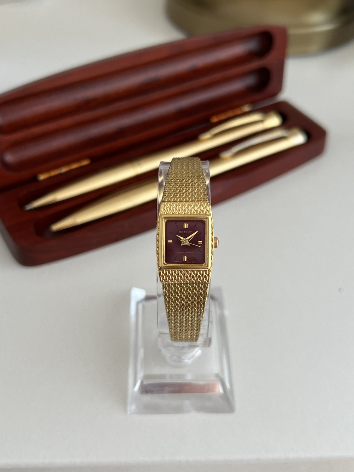 Orient women’s watch with a burgundy rectangular dial and textured gold tone mesh bracelet, shown front on.