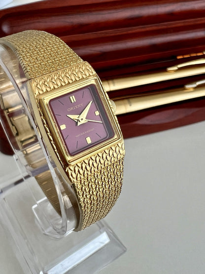 Close view of the Orient burgundy dial, gold tone hands and hour markers, with patterned case shoulders and mesh bracelet.