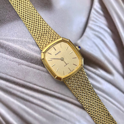 Rado women’s gold tone mesh bracelet watch with champagne dial, faceted case and blue cabochon crown worn on wrist.