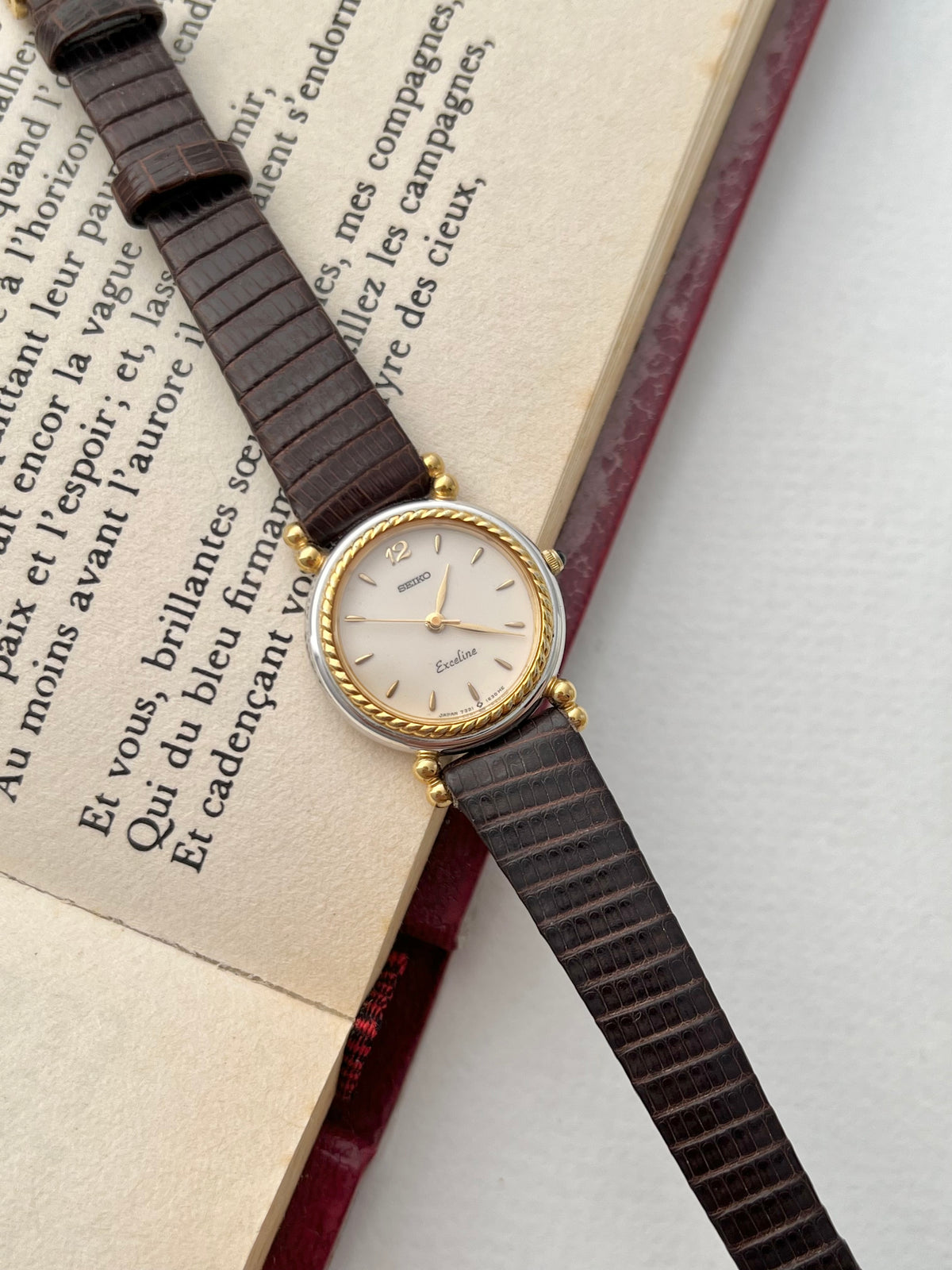 Seiko Two Toned Rope Bezel Woman's Watch — angled view of the pale dial, rope-textured gold bezel and dark leather strap on an open book.