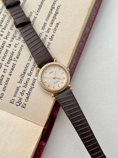 Seiko Two Toned Rope Bezel Woman's Watch — angled view of the pale dial, rope-textured gold bezel and dark leather strap on an open book.