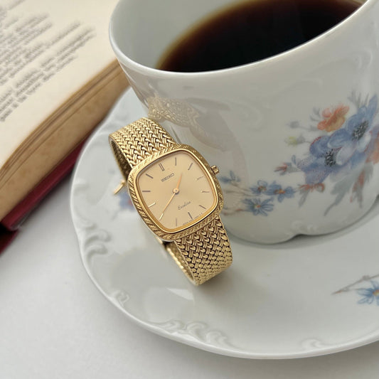 Seiko Exceline gold watch with diamond-set crown beside a cup and book.