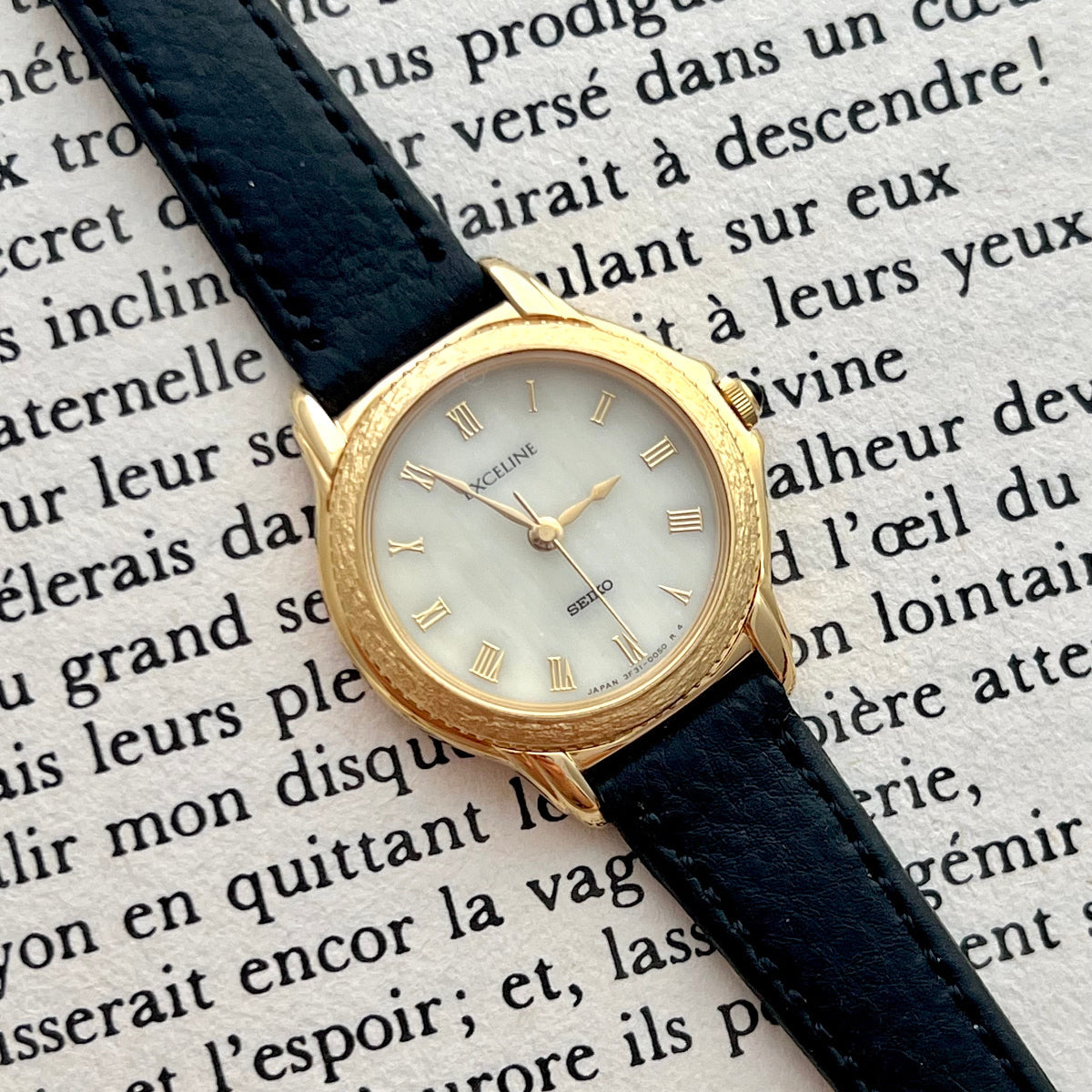 Seiko Exceline gold watch with black leather strap on a background of printed text.