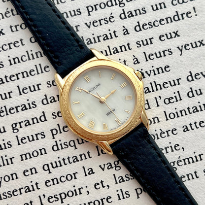 Seiko Exceline gold watch with black leather strap on a background of printed text.
