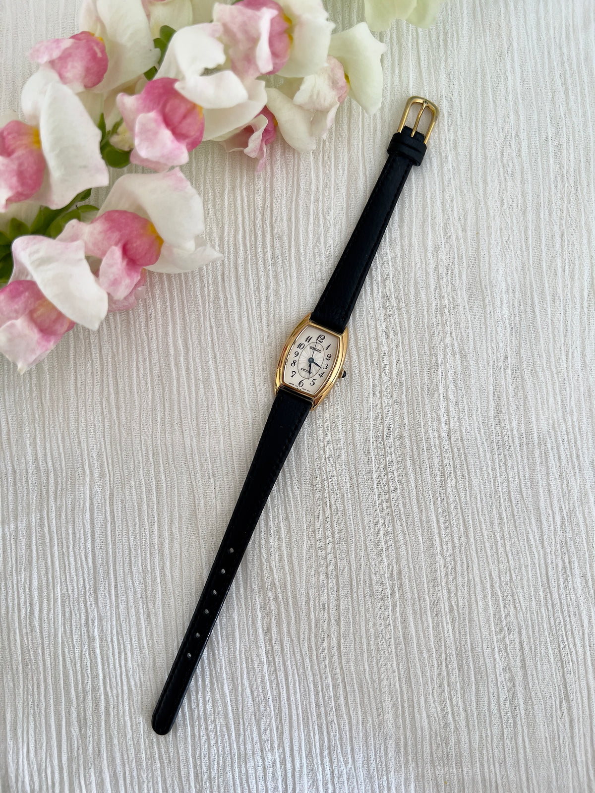 Seiko Exceline Gold Tonneau Women’s Watch with Arabic Dial — watch laid diagonally beside flowers.