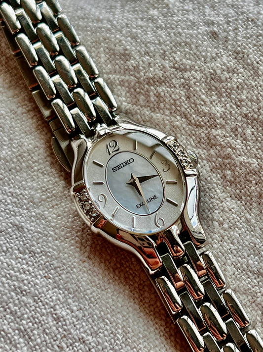 A Seiko Exceline Oval Diamond Bezel Woman's Watch with a mother-of-pearl dial and diamond accents on the bezel, displayed on a textured surface.
