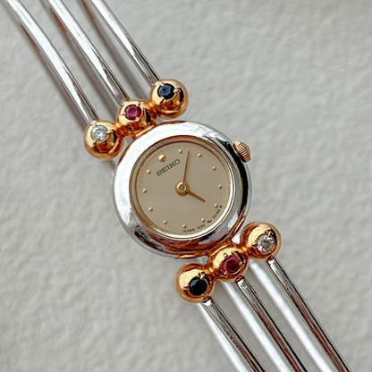 Seiko Tissé two-tone bangle watch with a champagne dial and ruby, sapphire and clear crystal accents; product image 1.