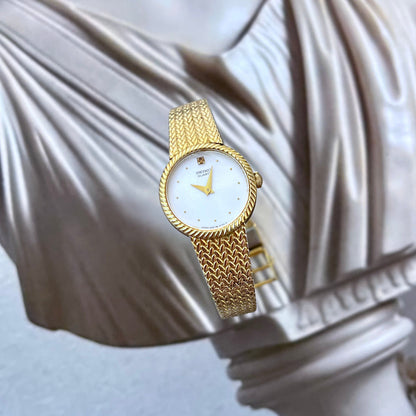 Seiko women’s gold tone watch with twisted rope bezel, white minimalist dial, crystal accent at twelve and woven bracelet styled against classical sculpture.