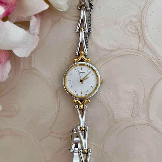 Seiko two-tone gold and silver watch displayed with an AVN pendant on a textured surface among flowers.