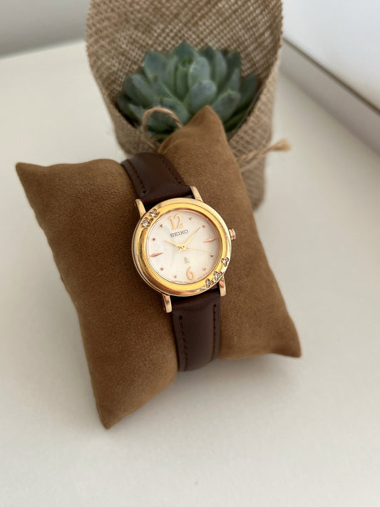 Seiko Lukia vintage gold-tone women's watch on brown velvet cushion, champagne numeral 12 and 6 dial and five gemstone accents on round bezel