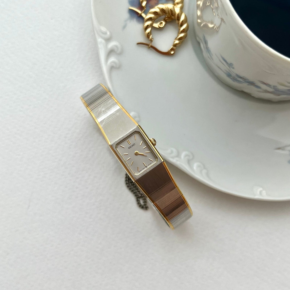 Seiko petite silver and gold bracelet watch on a white surface beside a cup and saucer.