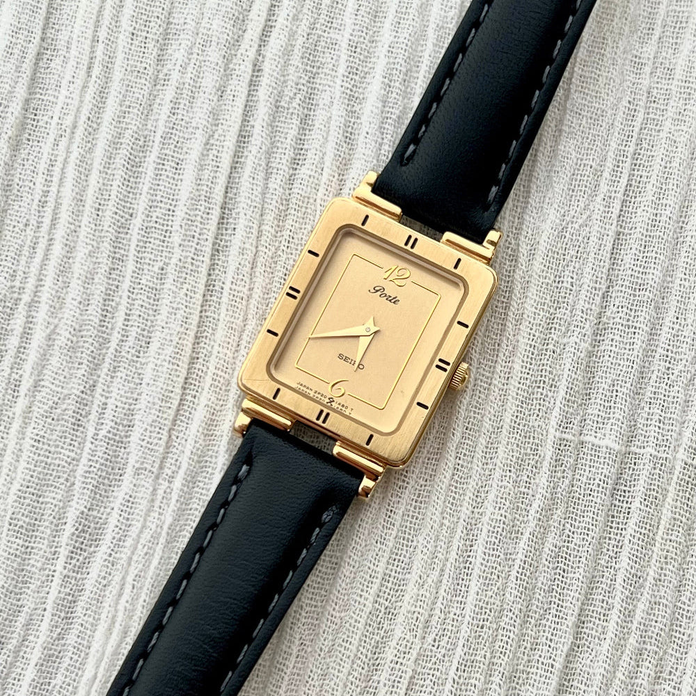Seiko Porte gold-tone Art Deco tank watch with black leather strap on a textured grey surface.
