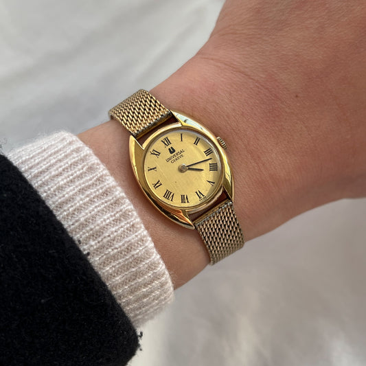 Universal Genève C case gold women’s Swiss watch with Roman numeral dial, vertical brushed finish and woven mesh bracelet worn on wrist.
