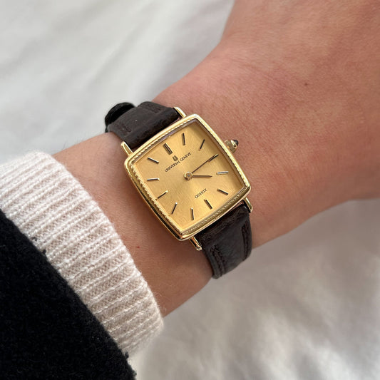 Universal Genève women’s Swiss watch with rectangular gold tone case, vertically brushed gold linen dial and brown textured leather strap worn on wrist.