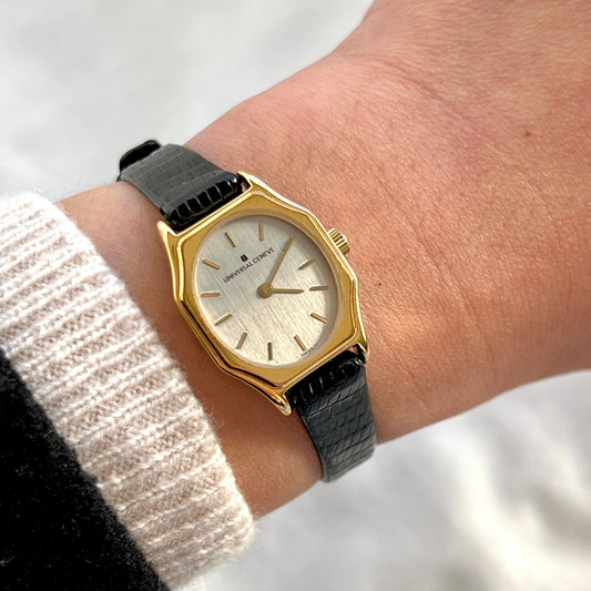 Universal Genève women’s Swiss watch with sculpted gold tone case, silver vertically brushed linen dial and black textured leather strap worn on wrist.