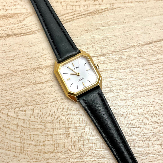 Vintage Rado Apis women's dress watch with a white dial, faceted gold-tone case and black leather strap, shown in a close front view.