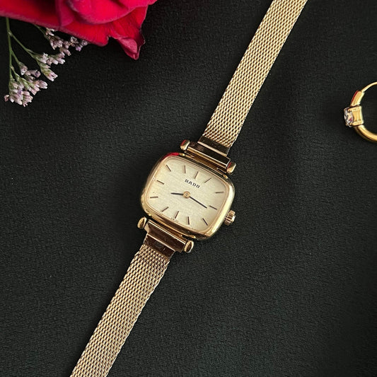 Vintage Rado cushion-square women's watch with a champagne dial and gold mesh bracelet, shown in a close front view with red roses.