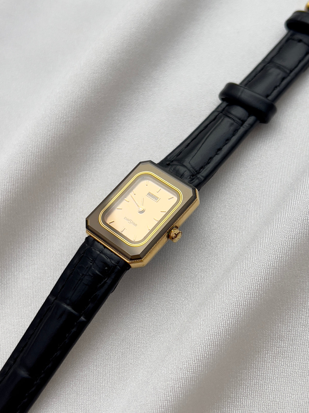 Vintage Rado DiaStar tank women's watch with a champagne dial and black leather strap, shown at an angle on white fabric.
