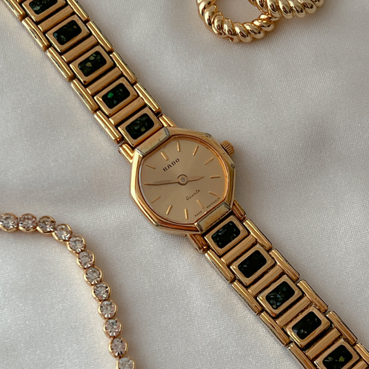 Vintage Rado octagonal women's watch with a gold dial and black abalone-inlay bracelet, styled with gold jewellery in a close flat lay.