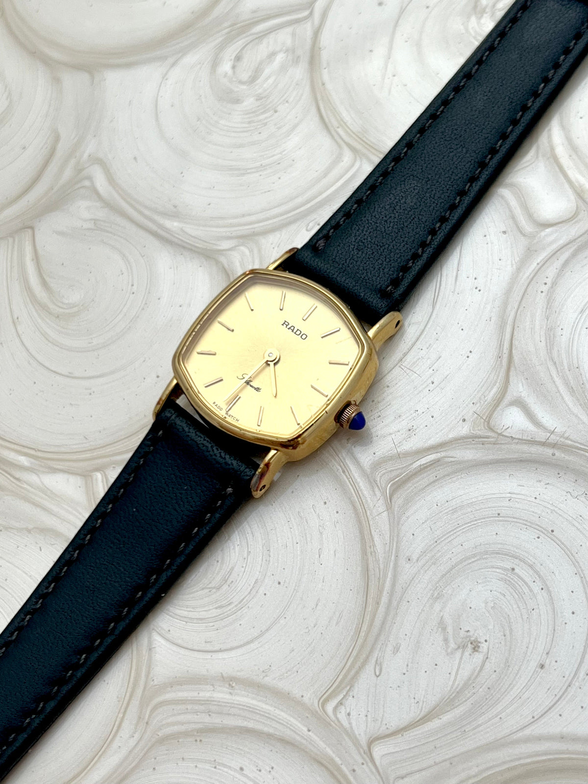 Vintage Rado Soft Octo Silhouette women's watch with a gold-tone case and black leather strap, shown from the side with its blue cabochon crown.