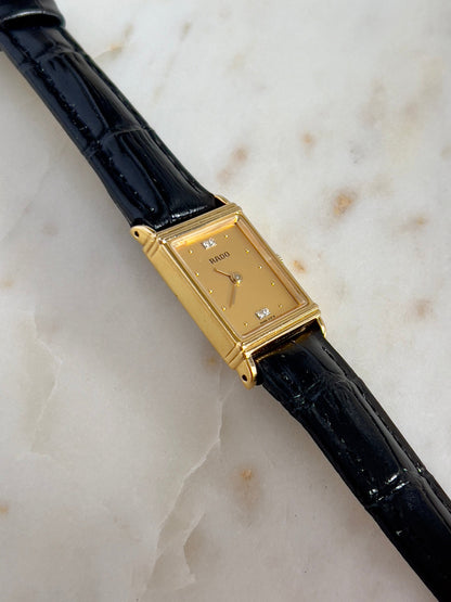 Vintage Rado tank women's watch with a gold dial, diamond accents and black leather strap, shown in an angled close-up.