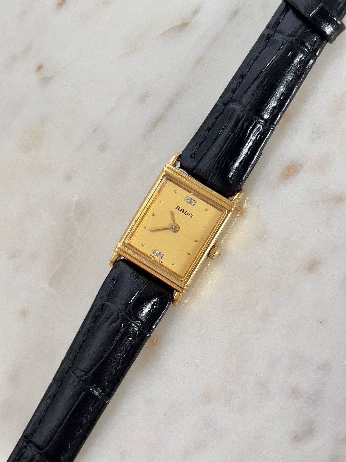 Vintage Rado tank women's watch with a gold dial, diamond accents and black leather strap, shown at an angle on marble.