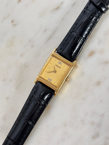 Vintage Rado tank women's watch with a gold dial, diamond accents and black leather strap, shown at an angle on marble.