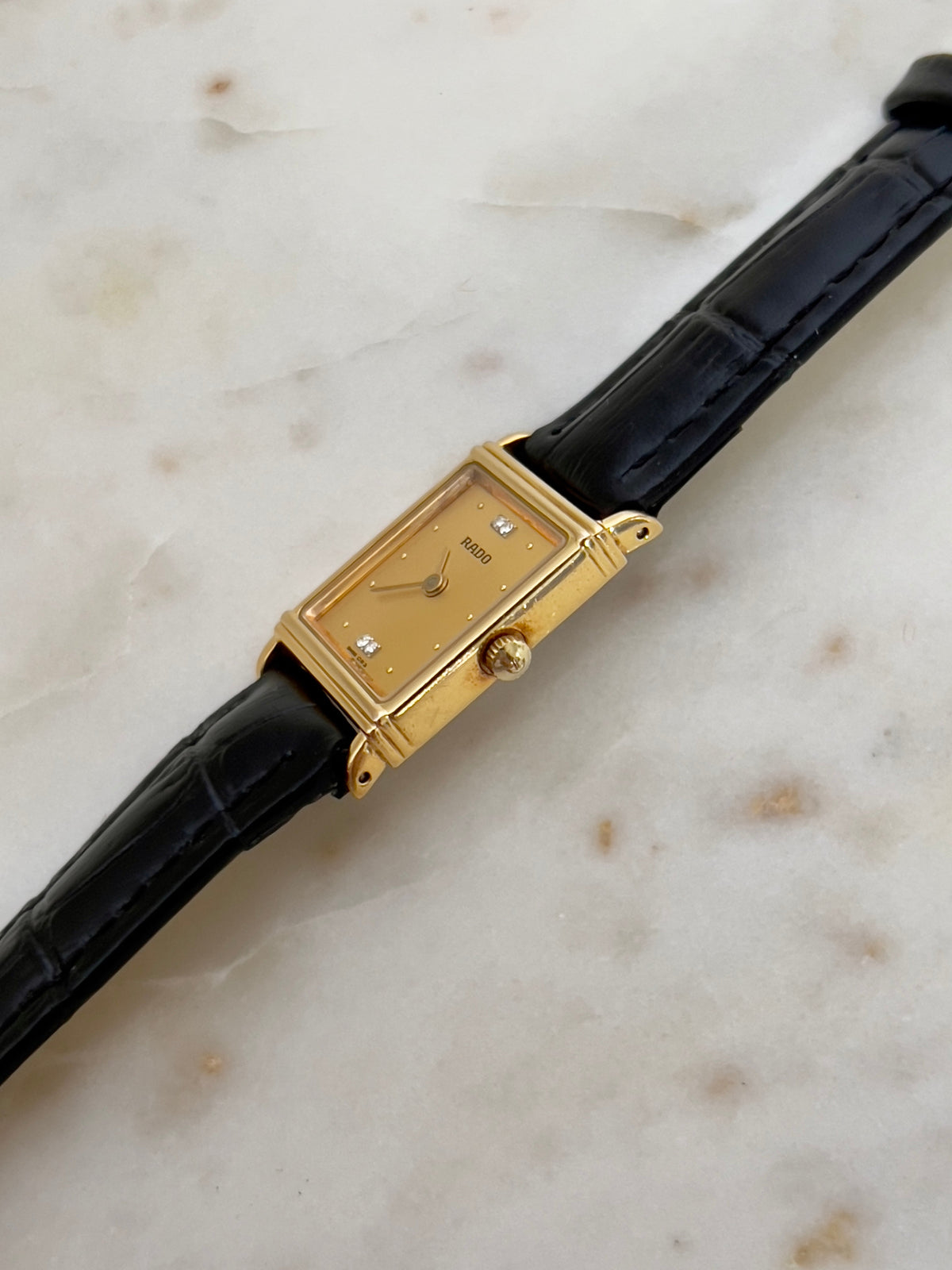 Vintage Rado tank women's watch with a gold dial, diamond accents and black leather strap, shown from the side.