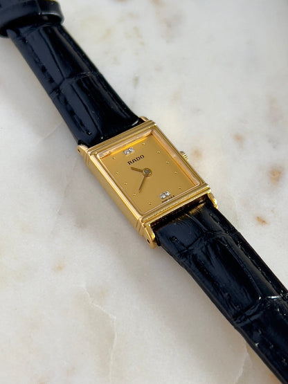 Vintage Rado tank women's watch with a gold dial, diamond accents and black leather strap, shown with the rectangular dial and stepped case.