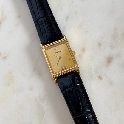 Vintage Rado tank women's watch with a gold dial, diamond accents and black leather strap, shown in a close front view.