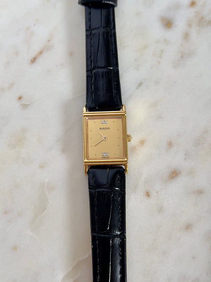 Vintage Rado tank women's watch with a gold dial, diamond accents and black leather strap, shown vertically in a front view.