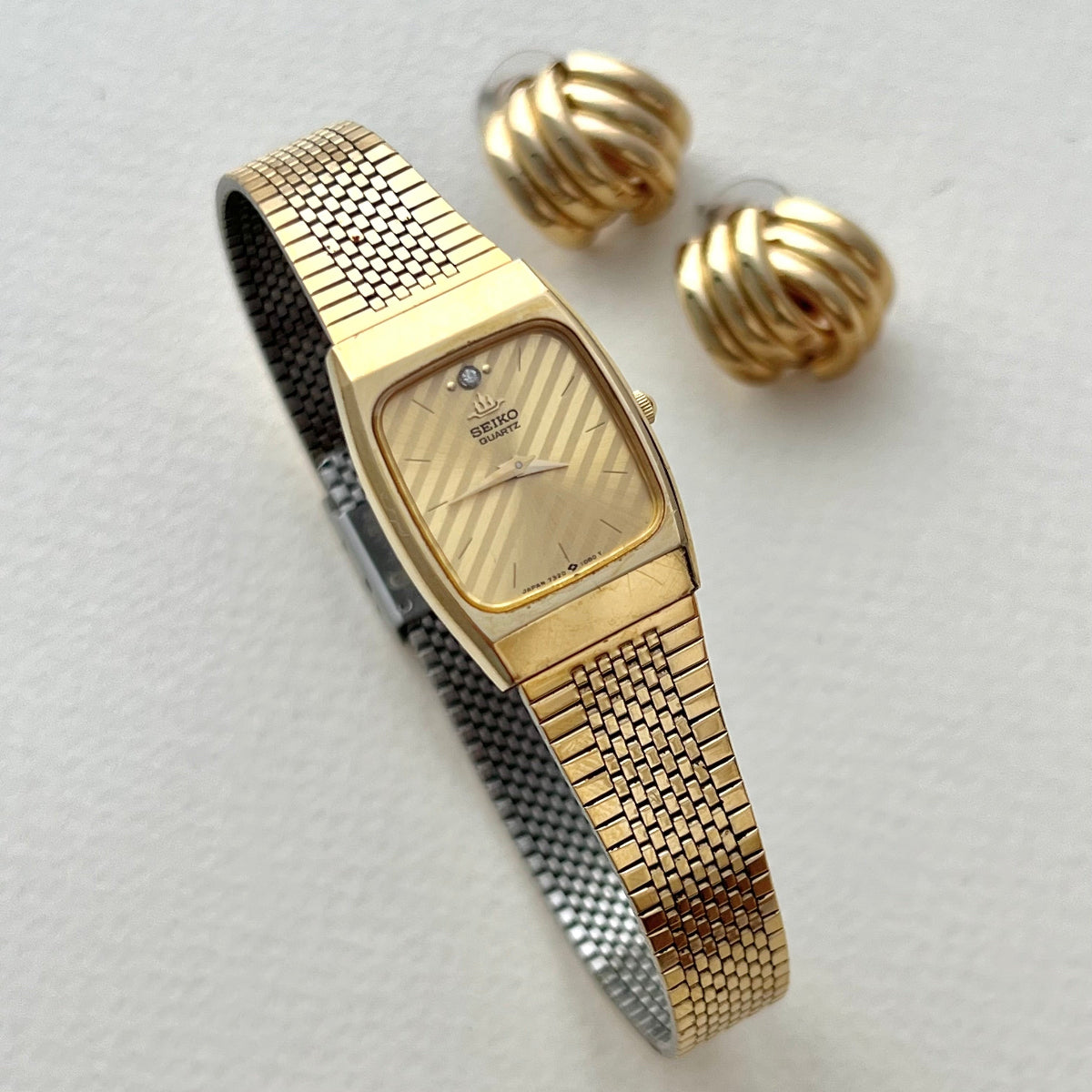 Vintage Seiko Quartz women’s watch with a rectangular gold-tone case, striped champagne dial, clear stone marker and integrated mesh bracelet styled beside gold earrings