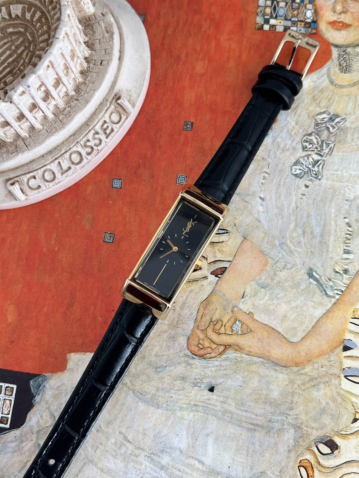 Yves Saint Laurent women’s watch with an elongated black dial, gold tone rectangular case and black leather strap in an art inspired still life.