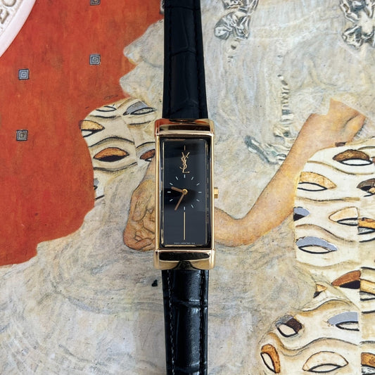 Straight front view of the Yves Saint Laurent elongated black dial watch with a gold YSL monogram and circular hour ring.