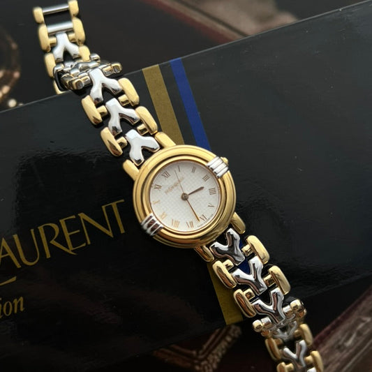 Close view of the Yves Saint Laurent textured silver white dial, gold tone Roman numerals and three hands.